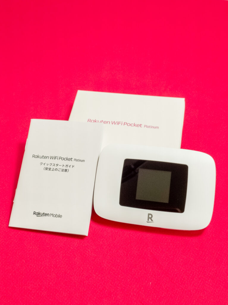 Rakuten WiFi Pocket Platinum本体とクイックスタートガイド。赤い背景の上に置かれた白いポータブルWiFiデバイスと付属のガイド。