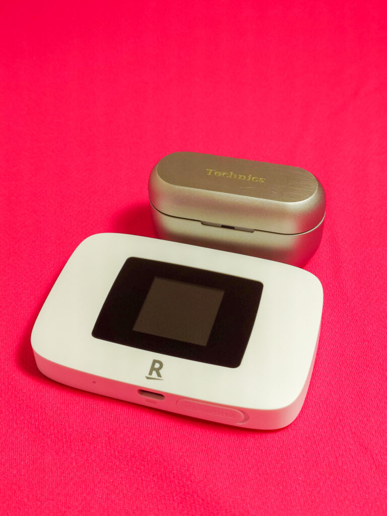 Rakuten WiFi Pocket PlatinumとTechnicsイヤホンケースが並べられた画像。赤い背景の上に置かれたデバイスのサイズ比較。」