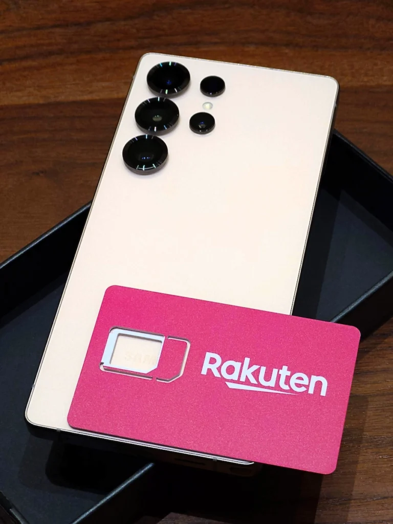 木目のテーブルに置かれたローズピンク色のGalaxy S25 Ultraと、楽天モバイルのピンク色SIMカード。背面には複数のカメラレンズが並んでいる。